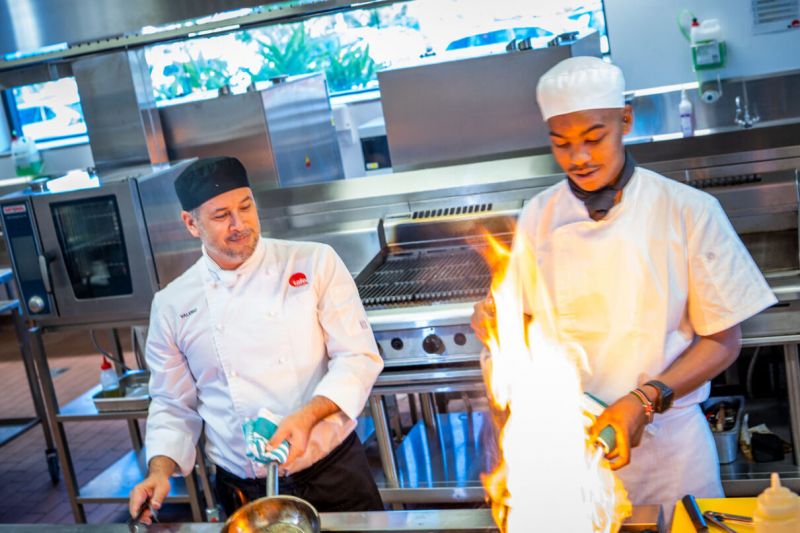 Two male chefs cooking in a kitchen, admiring a large flame that is blazing on a pan.